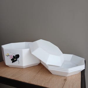 May include: White, geometric-shaped storage containers with lids. One container has a black and pink floral design. The containers are stacked on a wooden surface. The lids are off, revealing the inside of the containers.