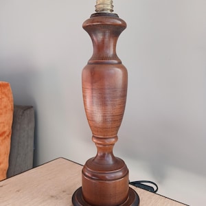 May include: A vintage wooden table lamp with a turned, urn-shaped body. The lamp has a dark brown finish and a brass bulb socket. The base is round and stepped. A black power cord extends from the base.