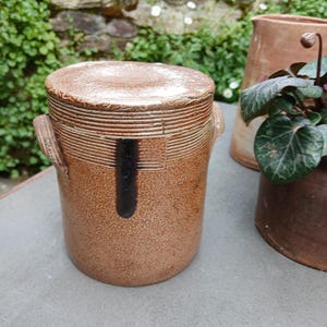 May include: A brown stoneware jar with a lid and two handles. The jar has a textured surface and a black stripe down the front.