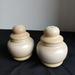Peut inclure: Deux salières et poivrières en céramique beiges, de forme arrondie et tachetée. Chaque récipient a un petit dessus arrondi percé de trous. Les salières et poivrières sont posées sur une surface sombre.