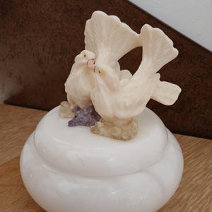 Könnte beinhalten: Weiße Keramik-Schmuckschatulle mit Deckel, gekrönt von einer Skulptur zweier sich küssender Tauben und einer dritten Taube im Hintergrund. Die Tauben sind cremefarben mit rosa Schnäbeln, und die Basis hat eine lila Traube.