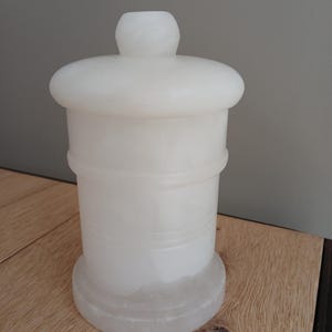 May include: A white alabaster jar with a lid. The jar has a cylindrical body with horizontal grooves and a rounded base. The lid is also rounded and has a small, cylindrical knob on top. The jar is sitting on a wooden surface.