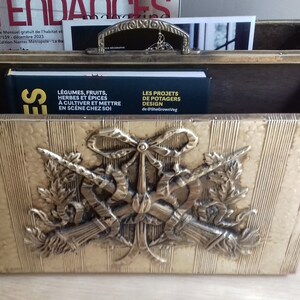 Puede incluir: Un revistero de metal dorado con un diseño decorativo de espadas cruzadas y un arco. El revistero está lleno de revistas y libros, incluyendo uno titulado "Les Projets de Potagers Design".