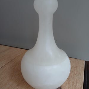 Könnte beinhalten: Eine weiße, undurchsichtige Glasvase mit bauchigem Boden und langem, schlankem Hals, der sich nach oben hin erweitert. Die Vase hat eine glatte, abgerundete Form und ein weiches, diffuses Aussehen. Die Vase steht auf einer Holzoberfläche.