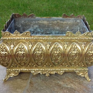 May include: A large, ornate brass planter with intricate floral designs. The planter has a rectangular shape with rounded edges and sits on four decorative feet.