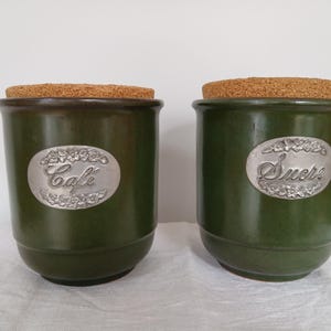 Peut inclure: Deux pots en céramique verte avec des couvercles en liège. Les pots ont des étiquettes argentées qui disent "Cafe" et "Sucre".