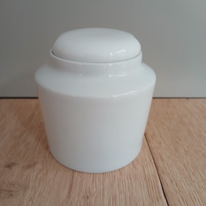 Puede incluir: Un tarro de cerámica blanco con tapa a juego. El tarro tiene un cuerpo redondeado y un acabado liso y brillante. La tapa encaja perfectamente en la parte superior, creando una estética limpia y minimalista. El tarro probablemente se utiliza para almacenamiento o fines decorativos.