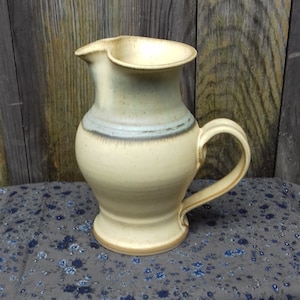 Puede incluir: Una jarra de cerámica de color crema con un esmalte azul y marrón. La jarra tiene una boca ancha y un asa.