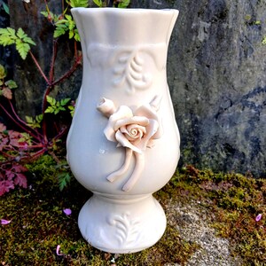 May include: A white ceramic vase with a delicate floral design and a raised rose accent. The vase has a fluted rim and a pedestal base.