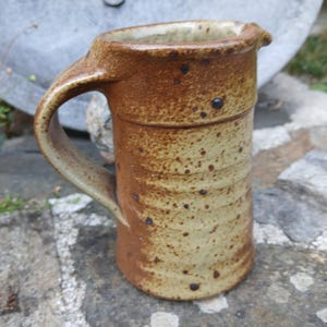 Puede incluir: Una jarra de cerámica hecha a mano con asa y boquilla. La jarra es de color beige claro con motas marrones y un borde y asa marrón más oscuro. La jarra está sobre una superficie de piedra.