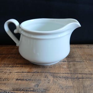 Op de afbeelding: Een witte keramische creamer met een gebogen handvat en een tuit. De creamer heeft een afgeronde body en een decoratieve band aan de bovenkant. De achtergrond is donker en de creamer staat op een houten oppervlak.