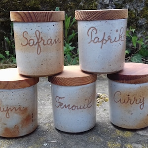 Peut inclure: Cinq pots à épices en céramique blanche avec des couvercles en bois. Les pots sont étiquetés avec les noms des épices en lettres brunes : Safran, Paprika, Thym, Fenouil et Curry.