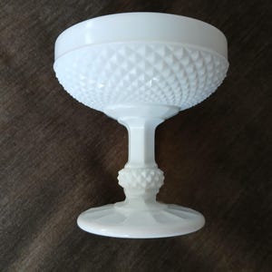 May include: A white milk glass pedestal bowl with a diamond pattern. The bowl is on a wooden surface.