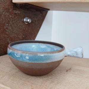 Peut inclure: Un bol à soupe en céramique fait à la main avec une poignée. Le bol présente un dégradé de couleurs, passant d'un intérieur bleu clair à un extérieur marron. La poignée est blanche. Le bol est posé sur une surface en bois.