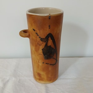 May include: A brown ceramic vase with a black abstract design and a small handle. The vase has a cylindrical shape and a slightly flared top.
