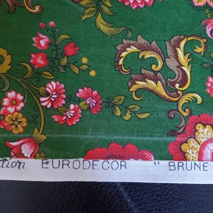 Puede incluir: Tela verde con un estampado floral con flores rojas y amarillas, y elementos decorativos marrones y amarillos. La tela tiene el texto "Création EURODECOR "BRUNE"" impreso en una tira blanca.