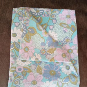 Può includere: Tessuto floreale piegato con un motivo ripetuto di grandi fiori blu, rosa, bianchi e oro su sfondo azzurro. Il tessuto è piegato, mostrando il motivo e la trama.