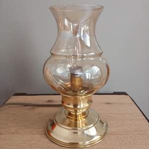 Könnte beinhalten: Eine goldene Tischlampe mit einem klaren Glasschirm. Der Schirm hat einen gewellten Rand und eine Glühbirne ist im Inneren sichtbar.