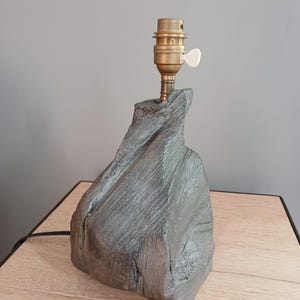 Op de afbeelding: Een grijze keramische tafellamp met een getextureerde, rotsachtige basis en een messing lampenkap. De lamp heeft een witte schakelaar aan de zijkant.