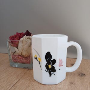 May include: A white octagonal ceramic mug featuring a graphic print of a large black flower with a yellow center, flanked by smaller yellow and pink floral motifs. The mug has a smooth finish and a curved handle on the right side of the body.