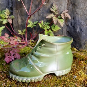 May include: A green ceramic boot planter with a white glaze on the inside. The boot has a lace-up design and a textured sole. The planter is sitting on a bed of moss.