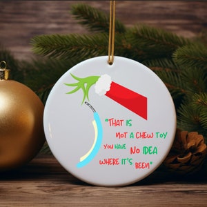 May include: A white ceramic Christmas ornament featuring a green Grinch hand holding a blue and white striped object. The ornament has the text "THAT IS NOT A CHEW TOY YOU HAVE NO IDEA WHERE IT'S BEEN" printed on it.
