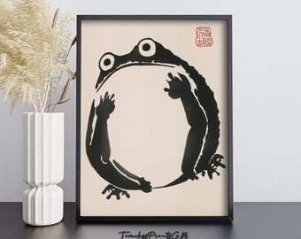May include: Black and white ink illustration of a frog in a circle, framed in a black frame. The image is on a beige background with a red stamp in the top right corner. The text "Timber Prints" is printed below the frame.