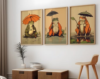 Op de afbeelding: Drie ingelijste illustraties van kikkers die oranje gewaden dragen en paraplu's vasthouden. De kikkers zitten op waterlelies in een vijver. De illustraties zijn in een minimalistische stijl met een witte achtergrond.