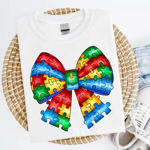 May include: White t-shirt with a colorful puzzle piece bow. The bow is made up of red, blue, green, and yellow puzzle pieces.