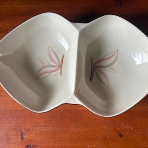 May include: A cream-colored ceramic divided serving dish with a symmetrical, elongated, leaf-like shape. Each of the two sections features a painted graphic of three stylized leaves in shades of soft pink, muted brown, and light gray.