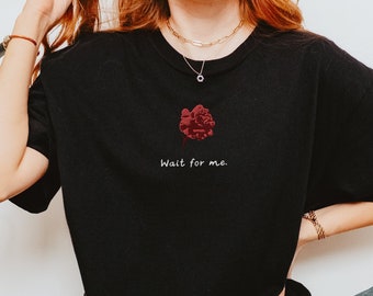 May include: A black t-shirt with a red rose graphic and the text "Wait for me." The model is wearing blue jeans and white sneakers.