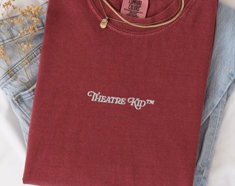 May include: A maroon t-shirt with the text "THEATRE KID™" embroidered on the front. The shirt is folded and placed on top of a pair of blue jeans.