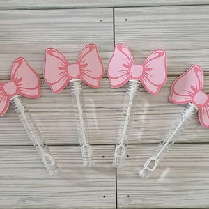 May include: Five pink bow-shaped bubble wands with clear plastic handles. The bows are attached to the handles with a small metal ring.