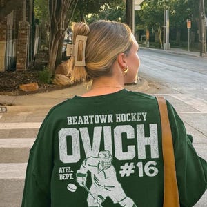 Puede incluir: Sudadera verde con un gráfico blanco de un jugador de hockey en la espalda. El gráfico dice "Beartown Hockey Ovich #16" y "Athl. Dept."