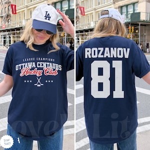 May include: Navy blue t-shirt featuring "Ottawa Centaurs Hockey Club" text and a hockey stick graphic. The back shows "Rozanov" and the number "81" in white. The shirt is worn with a white and blue baseball cap.