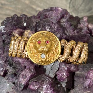 May include: A gold-toned bracelet featuring a lion's head with ruby eyes and a diamond in its mouth. The bracelet has intricate spiral detailing on the sides and is set against a purple, crystalline background.
