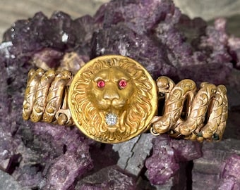 May include: A gold-toned bracelet featuring a lion's head with ruby eyes and a diamond in its mouth. The bracelet has intricate spiral detailing on the sides and is set against a purple, crystalline background.