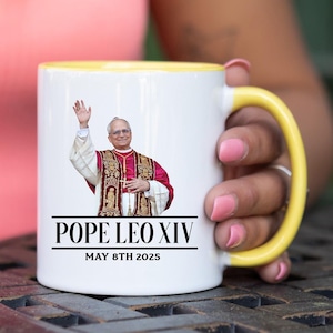 May include: White ceramic mug with a yellow interior and handle. The mug features an image of Pope Leo XIV and the text "POPE LEO XIV MAY 8TH 2025". The mug is held by a person.