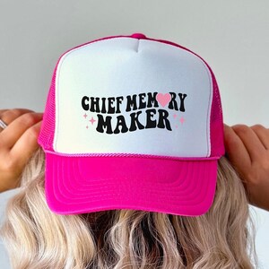 May include: A hot pink and white trucker hat with the text "CHIEF MEMORY MAKER" in black script. The hat has a mesh back and a curved brim. The text is decorated with small pink stars.