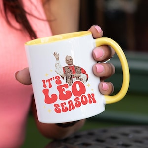 May include: White ceramic mug with a yellow rim. The mug features a graphic of a man in a red robe with the text "It's Leo Season" in red and yellow letters.