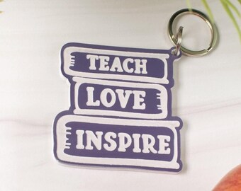 May include: A keychain featuring a stack of three books with the words "TEACH LOVE INSPIRE" in white lettering on a purple background. The keychain has a silver ring attached to the top. The background includes greenery and a partial view of a peach.