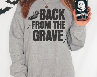 May include: A gray sweatshirt with the text "BACK FROM THE GRAVE" in black, along with a coffin graphic and bats. A black tumbler with the text "EVERYDAY IS HALLOWEEN" and a Michael Myers image is held in the hand.