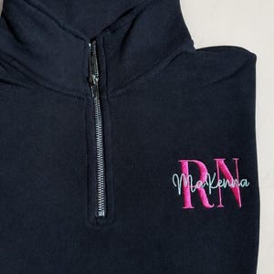 May include: A navy blue quarter-zip sweatshirt with a high collar. The sweatshirt features a silver zipper and embroidered text in pink and teal that reads "RN McKenna". The garment is folded neatly on a light-colored surface.