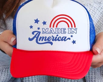May include: A trucker hat with a white front panel, blue mesh back, and a red brim. The front features a red rainbow graphic, blue stars, and the blue text "MADE IN America" printed in a combination of block and cursive fonts on the white panel.