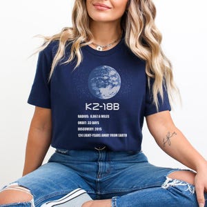 May include: A navy blue t-shirt with a graphic of a planet and the text "K2-18B". The text below the graphic reads "Radius: 8,867.6 miles, Orbit: 33 days, Discovery: 2015, 124 light-years away from Earth."