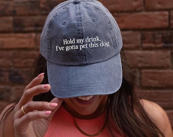 May include: A washed-out navy blue baseball cap featuring the white embroidered text "Hold my drink, I've gotta pet this dog" centered on the front panel. The hat has a structured crown, a curved brim, and stitched eyelets for ventilation.