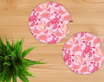 Puede incluir: Dos posavasos redondos de color rosa con un patrón repetido de tortugas marinas, conchas y cangrejos de color rosa y naranja.