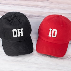 May include: A black baseball cap with the white text OH and a red baseball cap with the white text IO. Both caps feature a structured crown, a curved brim, and a button on top, displaying bold block lettering centered on the front panels.