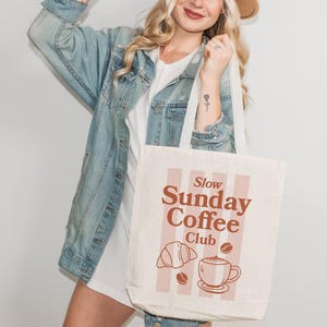 May include: Cream-colored tote bag with the text "Slow Sunday Coffee Club" in brown. The design includes a croissant, coffee beans, and a coffee cup. The bag has white straps and is held by a person.