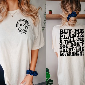 May include: A white t-shirt with a black graphic on the front that says "Buy Me Plants" with a dripping smiley face. The back of the shirt has black text that says "Buy Me Plants & Tell Me You Don't Trust The Government."